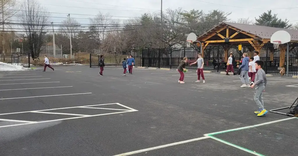 Children playing basketball on an outdoor court in winter, with a wooden pavilion and spectators in the background.