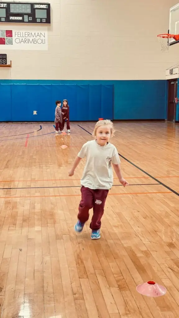 A young child runs toward the camera on a gymnasium hardwood floor, with a basketball hoop and blue padded wall in the background.