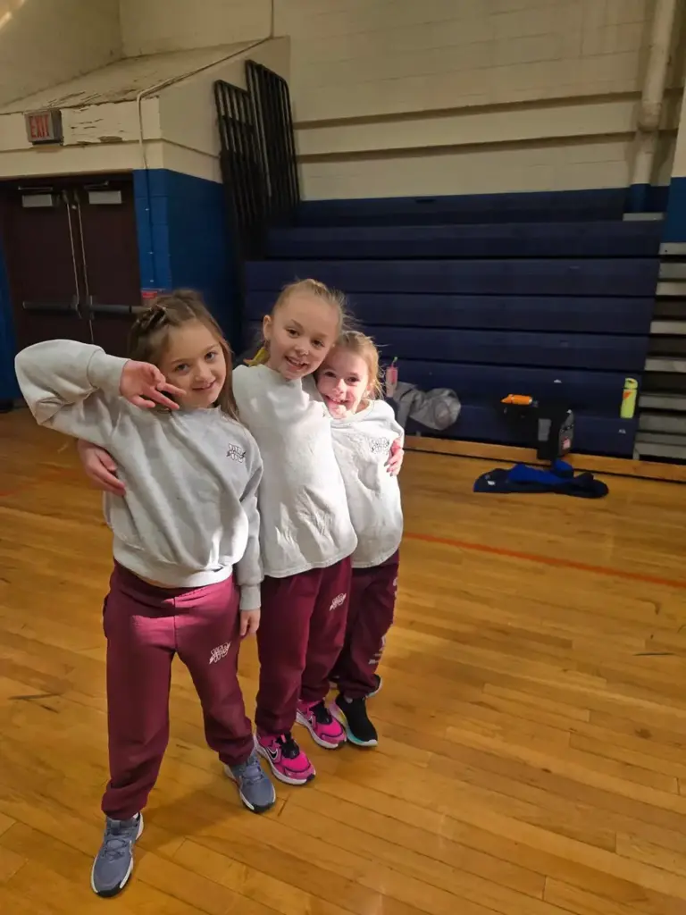 Three young girls in matching white sweatshirts and pink leggings pose together in a gymnasium with a wooden floor.