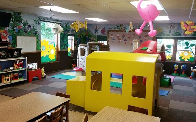 Bright preschool classroom with a yellow playhouse, kid-sized tables, shelves, and colorful wall decorations.