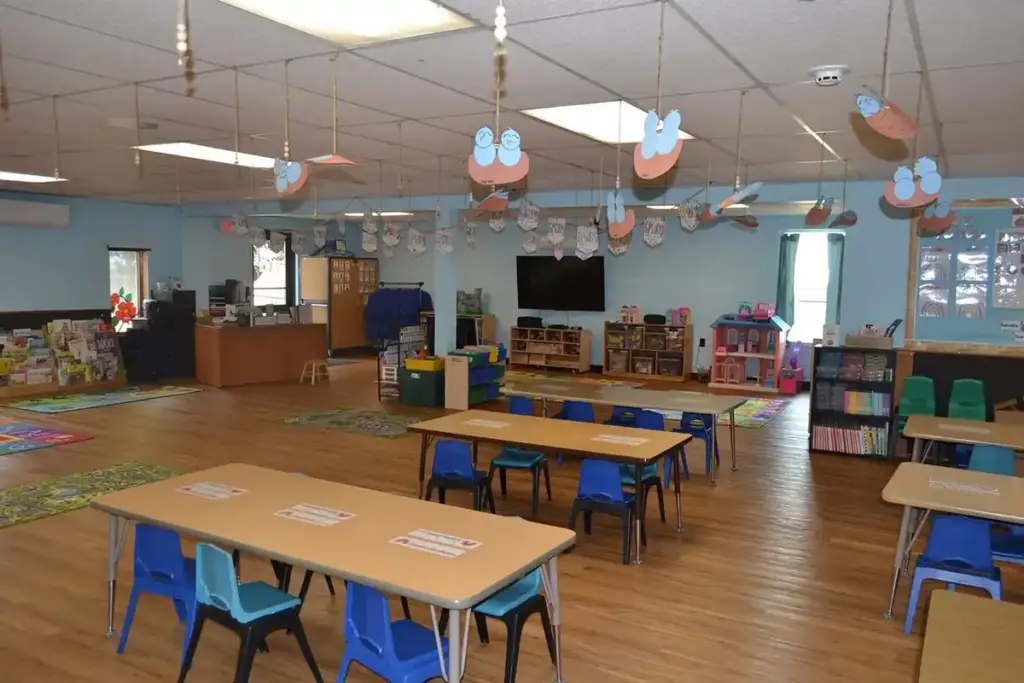 Bright preschool classroom with several low wooden tables, blue chairs, shelves of toys, and colorful decorations.