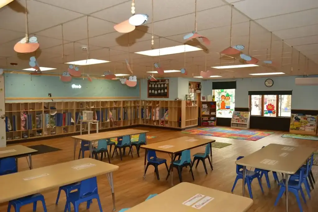 Bright kindergarten classroom with several tables and blue chairs, cubbies lining the walls, and a reading corner near a display screen.