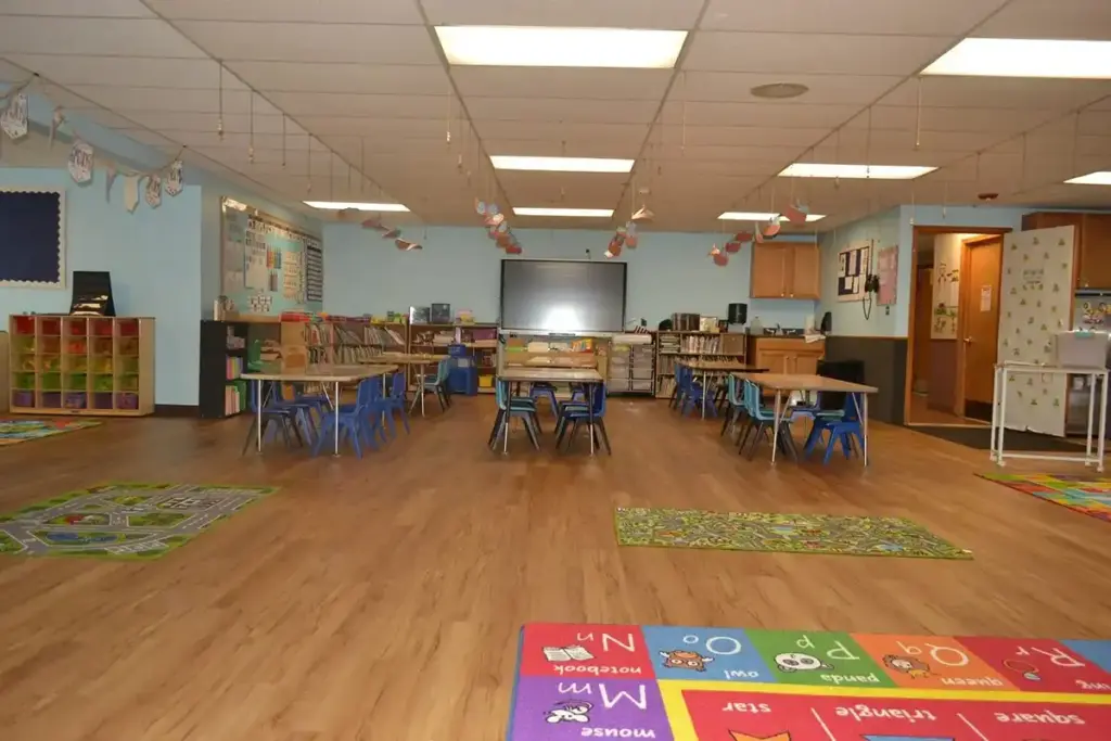 Brightly lit classroom with small tables and chairs arranged in the center, colorful rugs on a wooden floor, and shelves of supplies along the walls.
