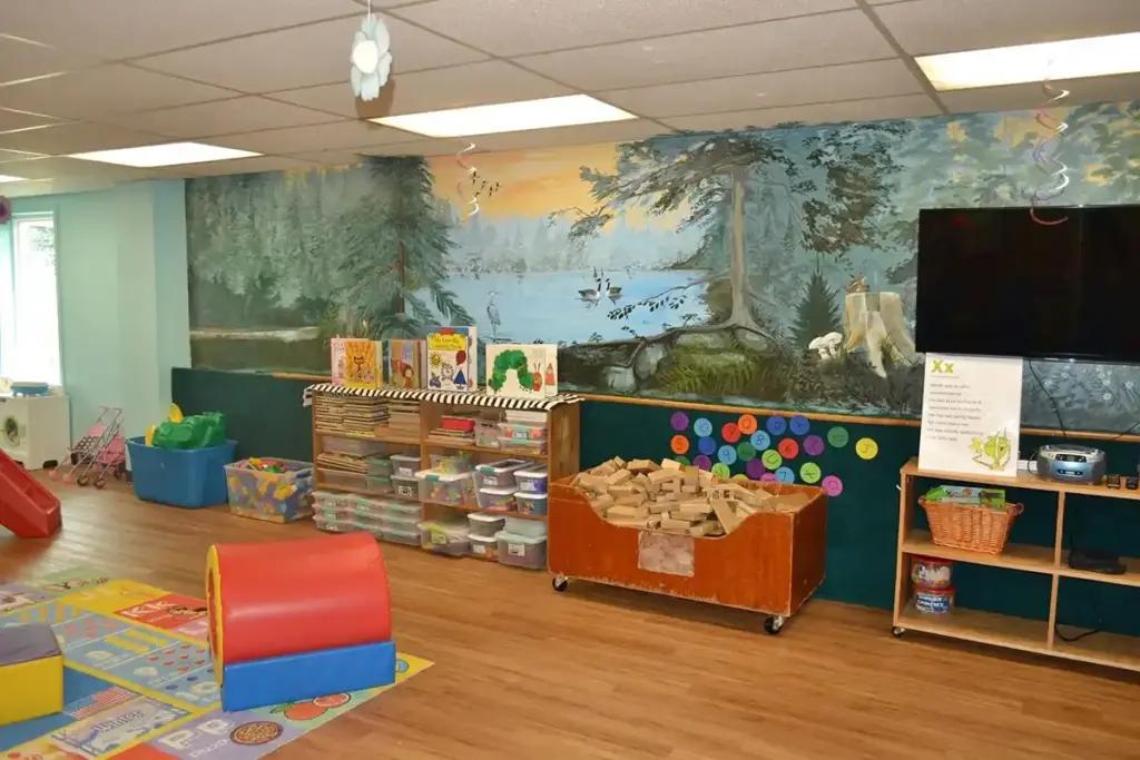 Bright preschool classroom with low shelves of toys, colorful bins, a soft play rug, and a tree mural on the wall.