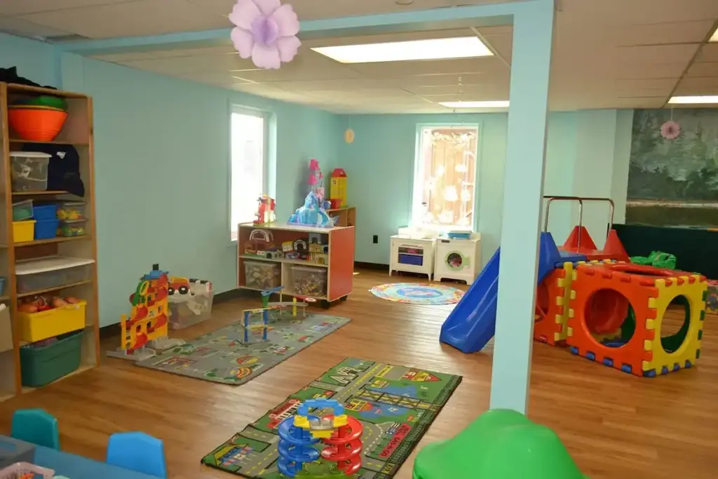 Colorful children's playroom with toy bins, a soft mat area, a small kitchen, a blue slide, and a climbing structure.