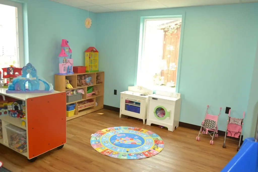 Bright children's playroom with toy shelves, a round colorful rug, a white play kitchen, and small pink chairs against light blue walls.