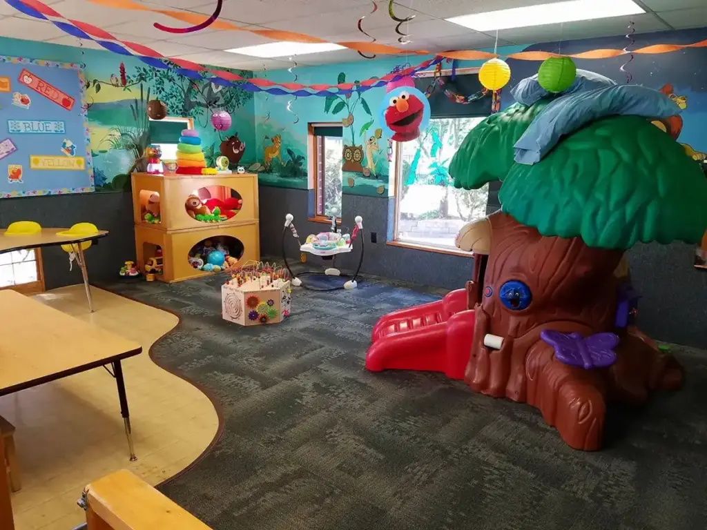 A bright children's playroom with a large tree-shaped play structure, a small red slide, toy shelves, and colorful wall murals.