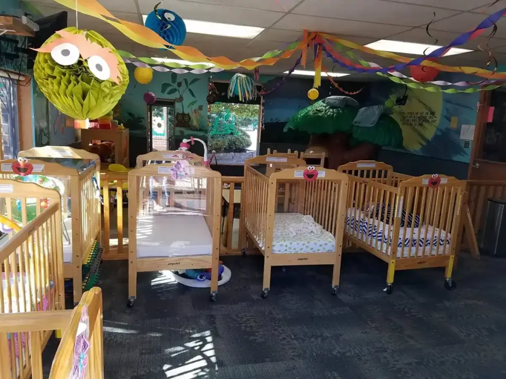 A children's daycare room with several wooden cribs arranged in a row, and colorful streamers and large green monster decorations hanging from the ceiling.