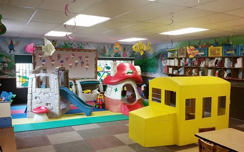 Colorful indoor children's play area with soft-play structures, including a yellow block train, a pink playhouse, a slide, and shelves of books along the wall.